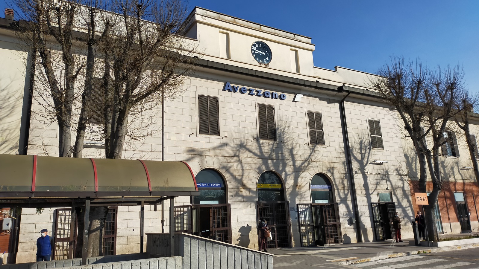 The street frontage of Avezzano railway station in Abruzzo, with the station name above the entrance arches and a taxi sign at the kerb