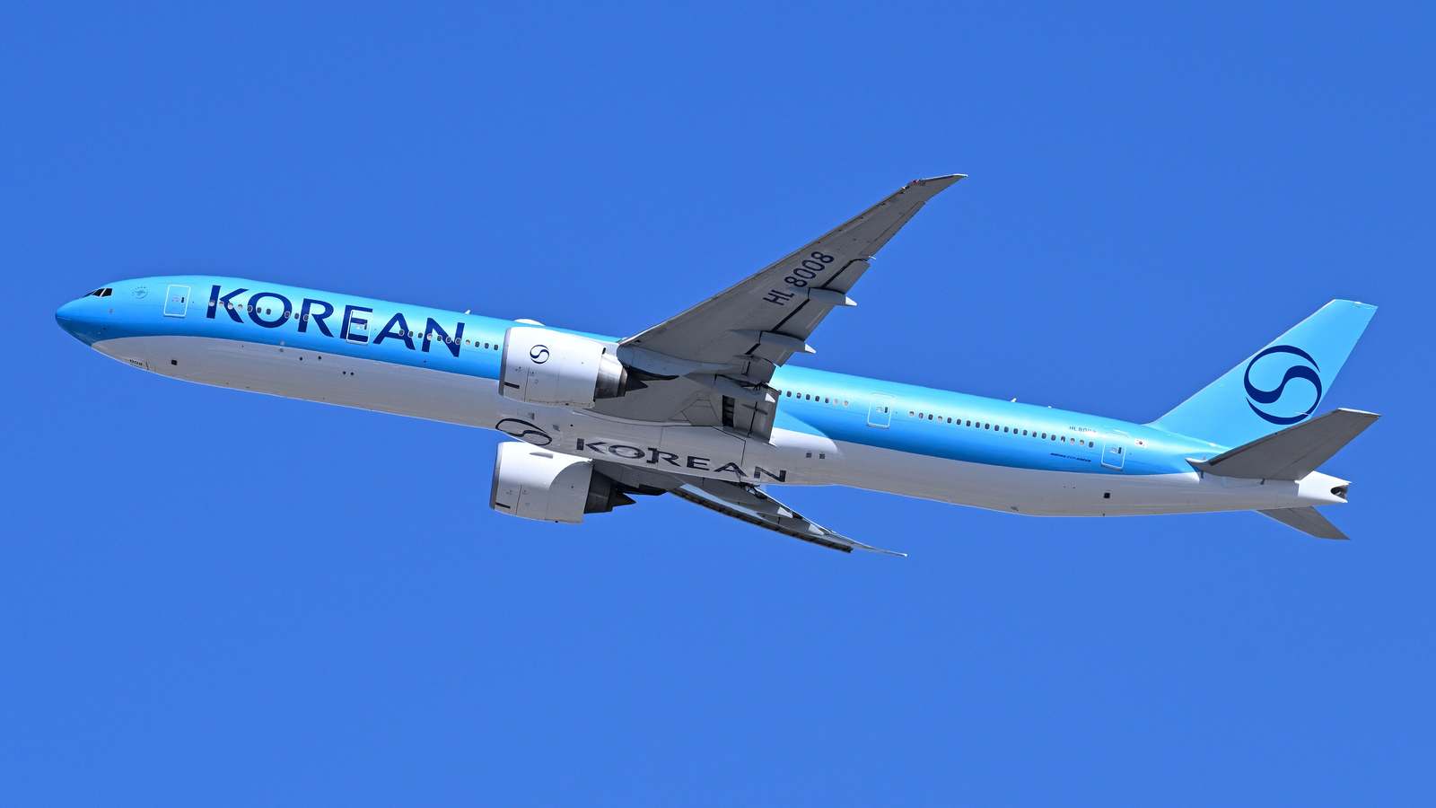 A Korean Air Boeing 777-300ER in blue and white livery climbing against a clear blue sky after departing Boston