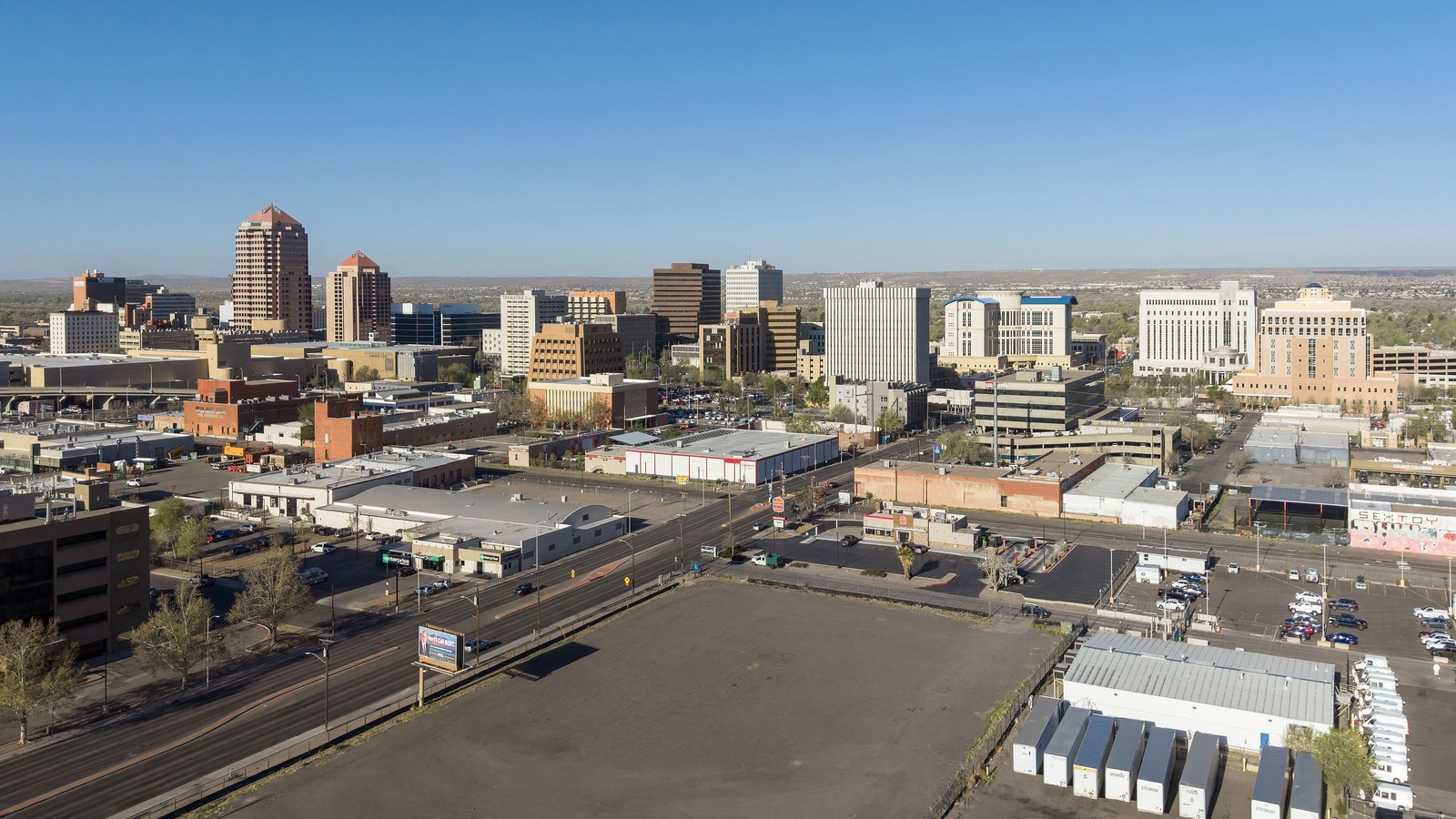 Downtown Albuquerque from the air on a clear day, mid rise office towers and hotels above low warehouses and surface parking, with the New Mexico mesa on the horizon.