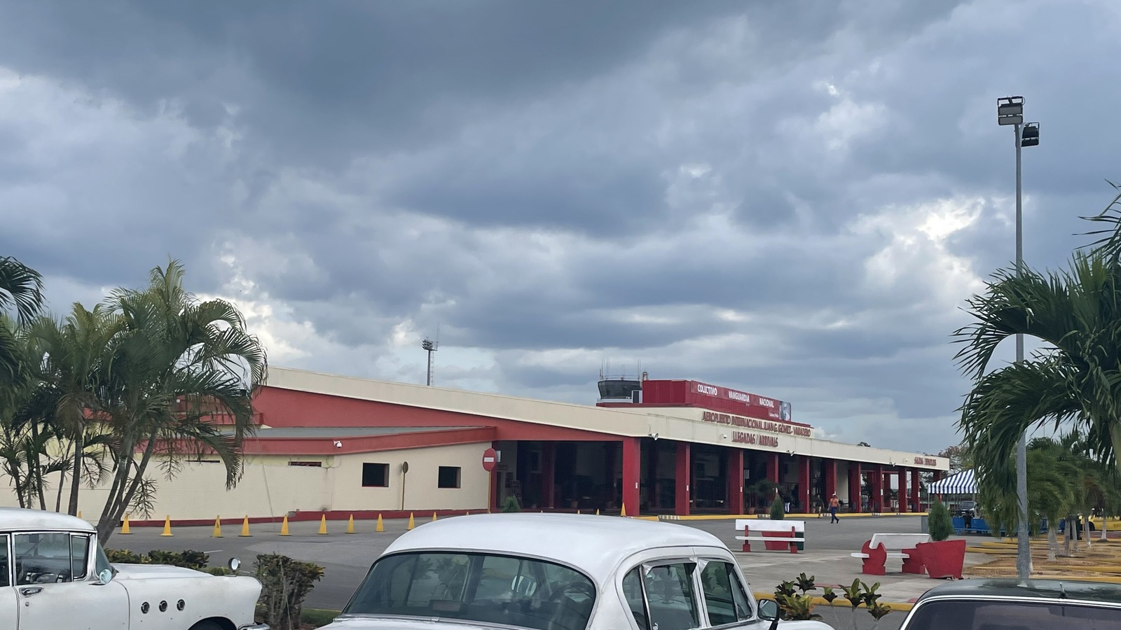 The red and cream arrivals terminal at Varadero's Juan Gualberto Gómez International Airport in Cuba under a grey sky, with palm trees and classic cars in front.