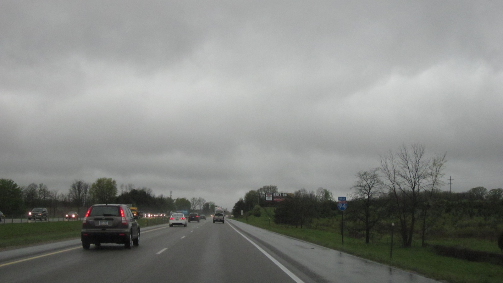 Wet Interstate 94 in Michigan with a posted I-94 shield and traffic in the travel lanes under an overcast sky.