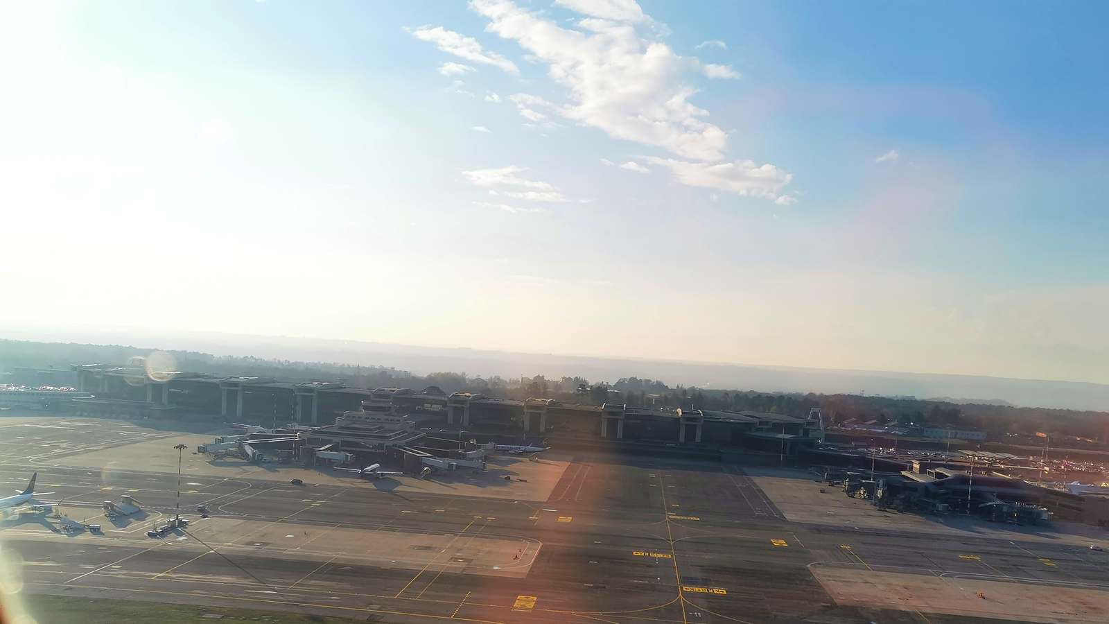 Milan Malpensa Terminal 1 seen from an aircraft window, with the long terminal building, jet bridges and parked aircraft on the apron in low sun.