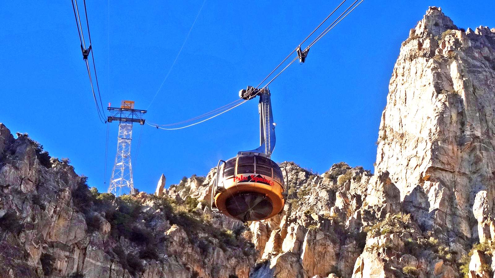 Rotating Palm Springs Aerial Tramway car on its cables below a steel tower, climbing past sunlit granite cliffs under a deep blue sky.
