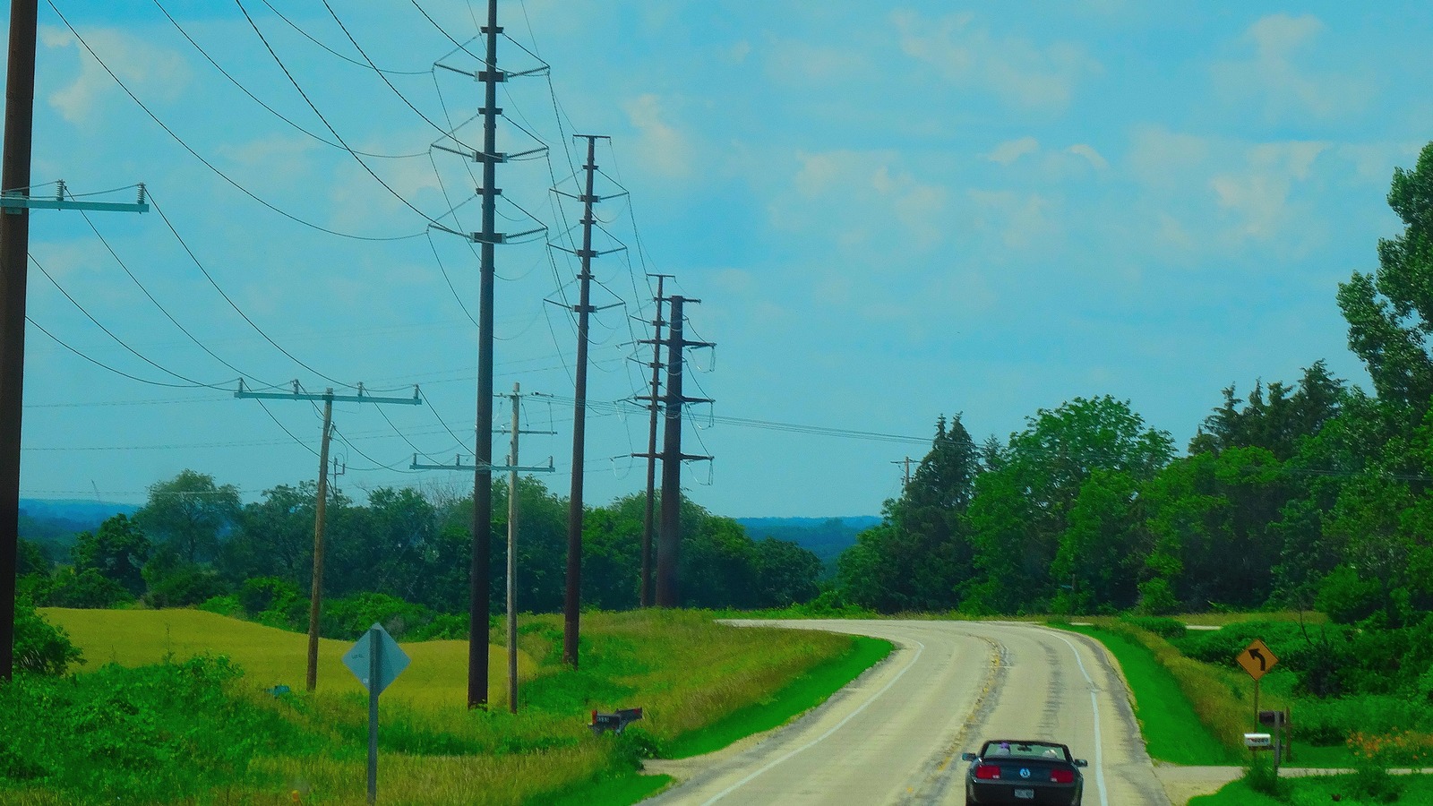 Daylight view of rural County Trunk Highway M curving past fields with a car on the road