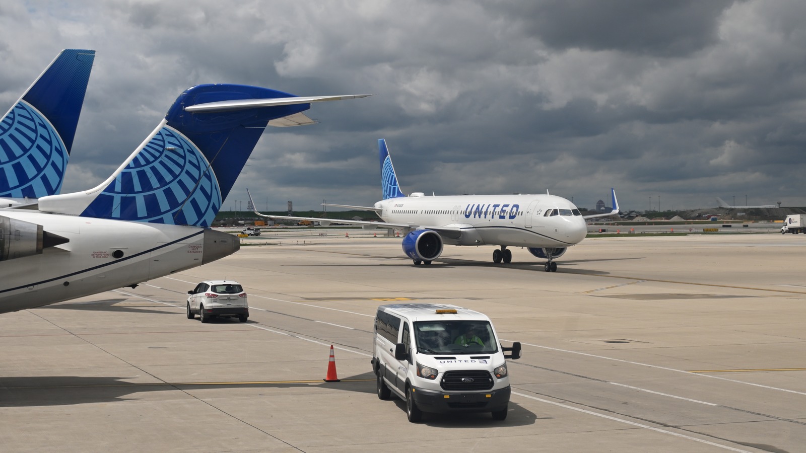 A United Airlines Airbus A321neo in the current blue livery taxis across the apron at Chicago O'Hare, with two other United tail fins and a United ground van in the foreground under heavy clouds.