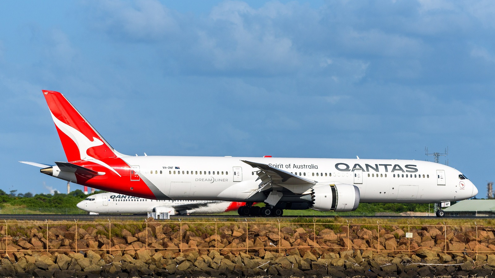 A Qantas Boeing 787-9 on the runway at Sydney Airport with a second Qantas jet behind and a rock seawall in the foreground.