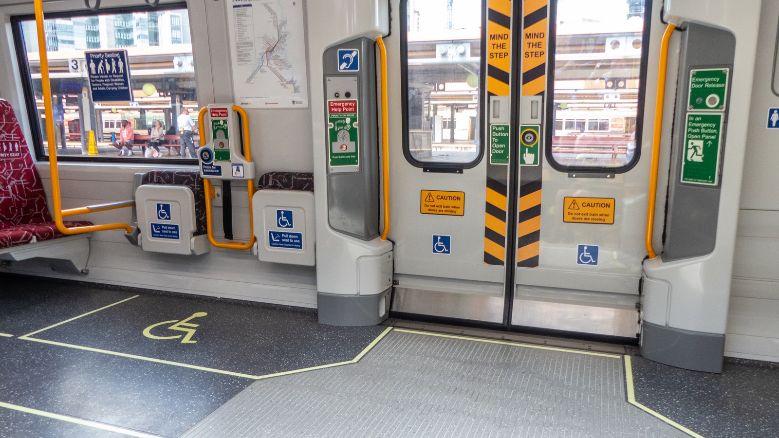 Interior door vestibule of a refurbished Queensland Rail New Generation Rollingstock train, showing yellow grab poles, wheelchair priority space, "mind the step" door markings and a route map, photographed at Roma Street station in Brisbane.