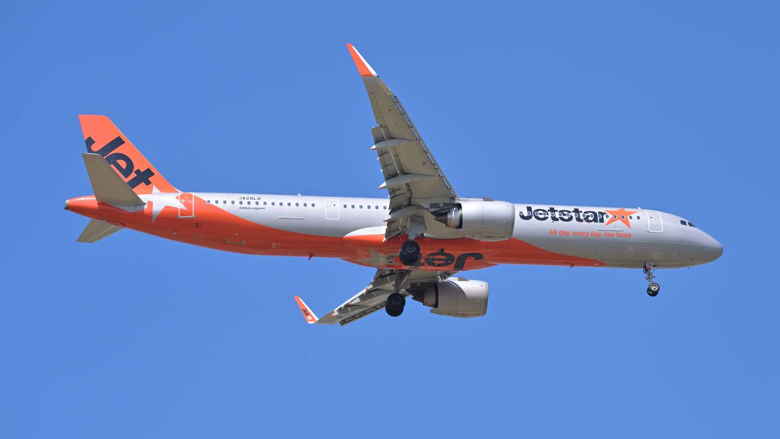 Jetstar Japan Airbus A321neo in silver and orange livery flying against a clear blue sky with landing gear down.
