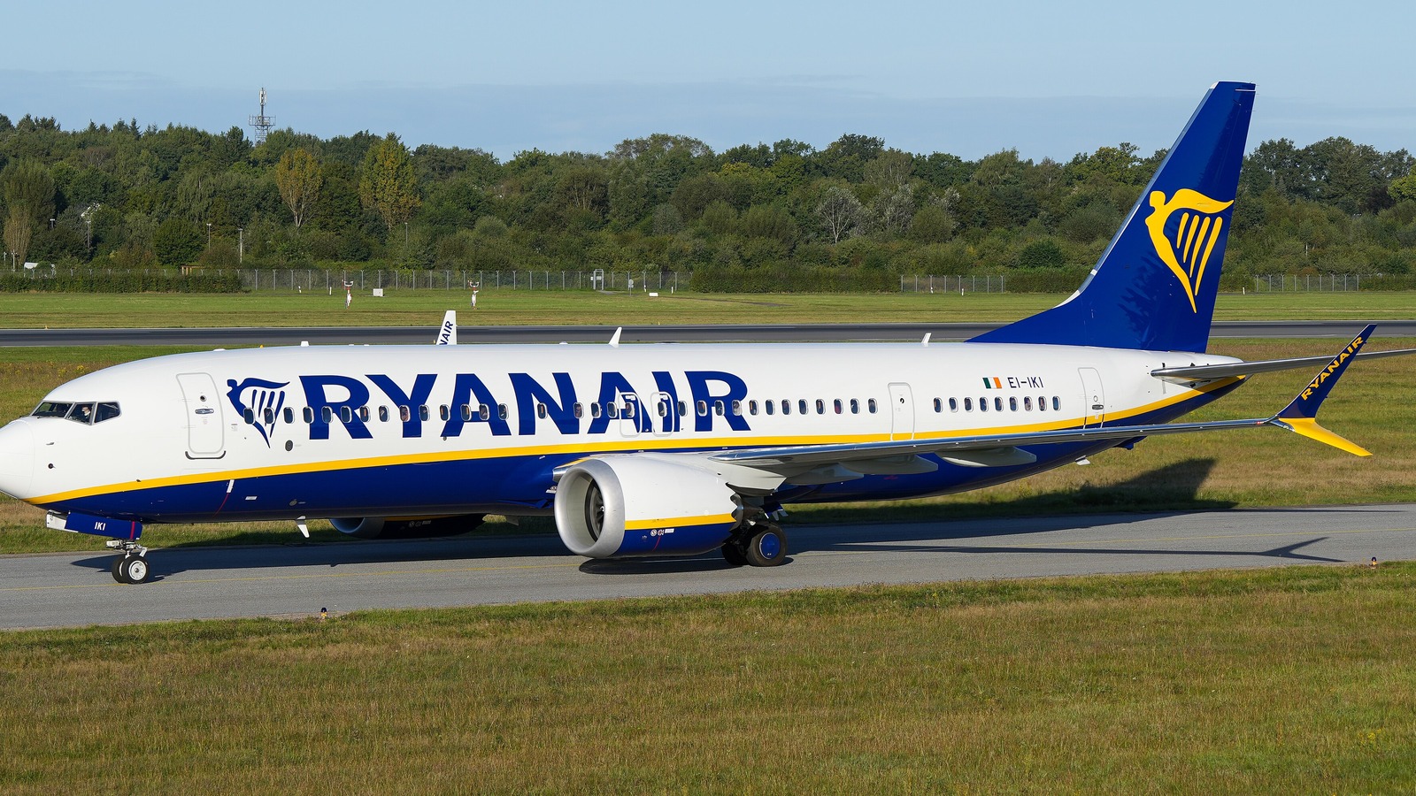 A white and blue Ryanair Boeing 737-8200 taxiing past green trees under a blue sky.
