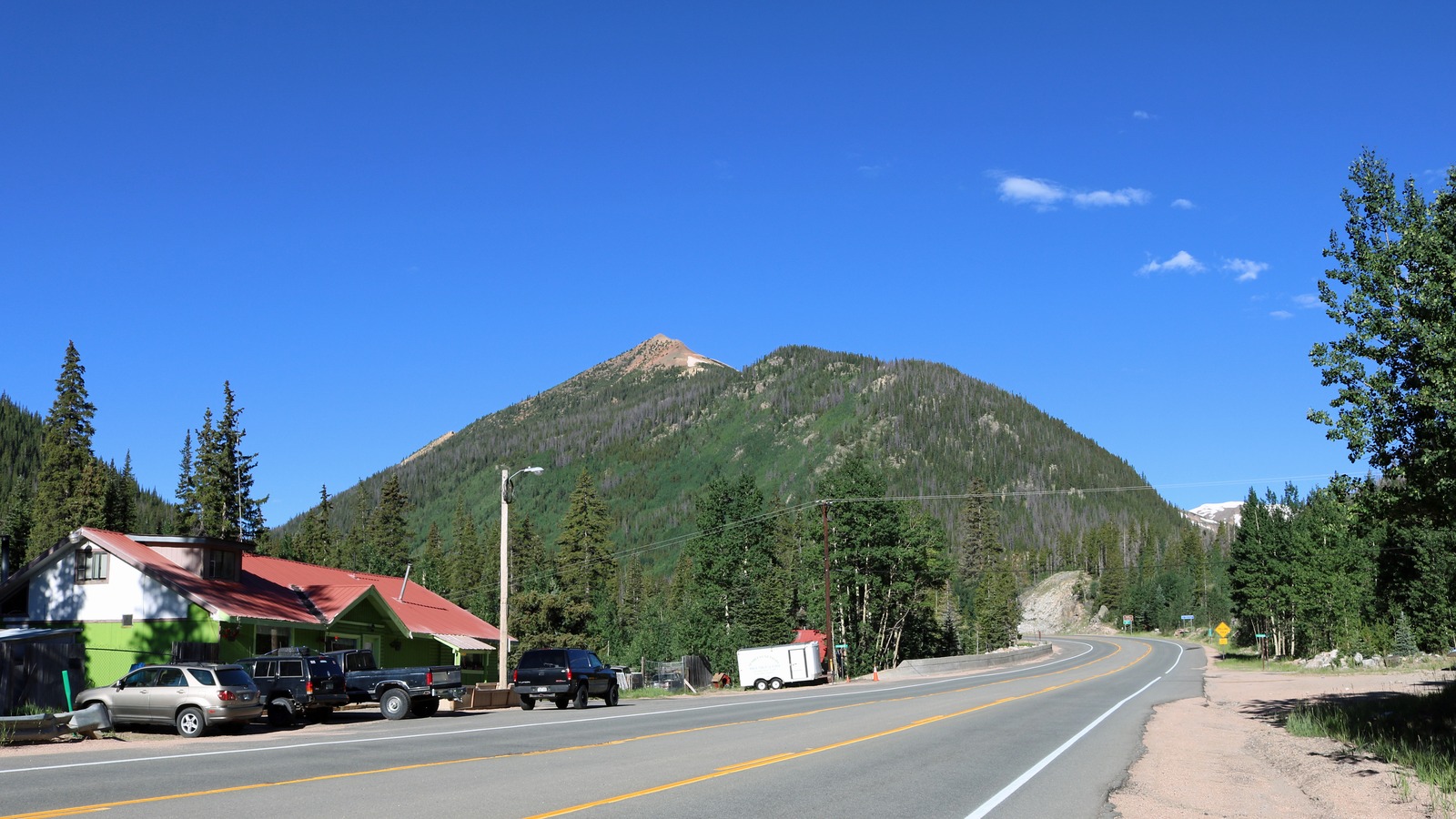 US 40 climbing towards Berthoud Pass at Berthoud Falls in Colorado, a two lane highway with a double yellow centre line curving right between forested slopes, with a rock cut visible beside the road and a snow patch on a distant peak
