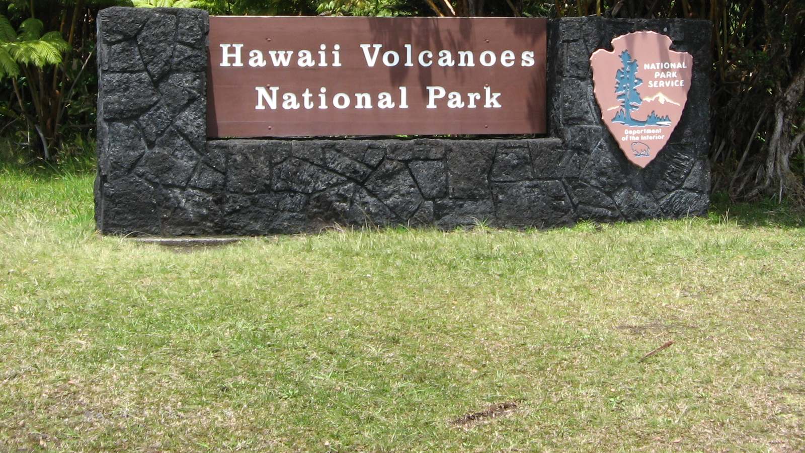 Hawaii Volcanoes National Park entrance sign on a lava rock wall with the National Park Service arrowhead, backed by ferns.