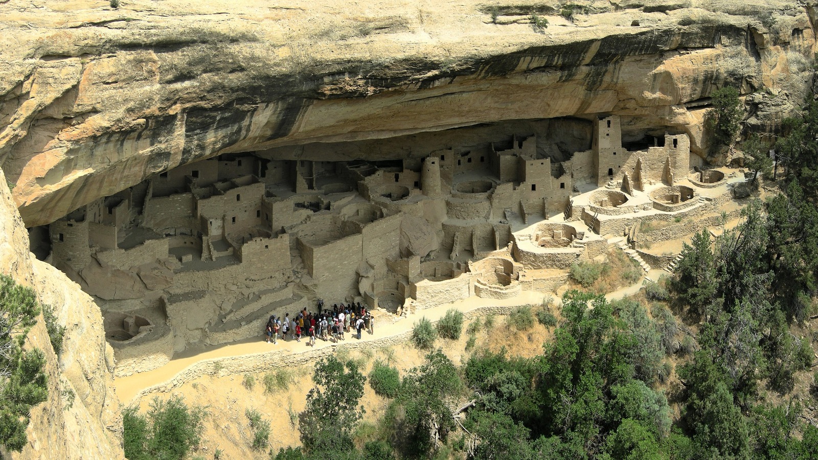 Cliff Palace seen from the overlook opposite, its sandstone towers and round kivas set under a long rock alcove, with a ranger led tour group standing together on the path in front of the dwelling.