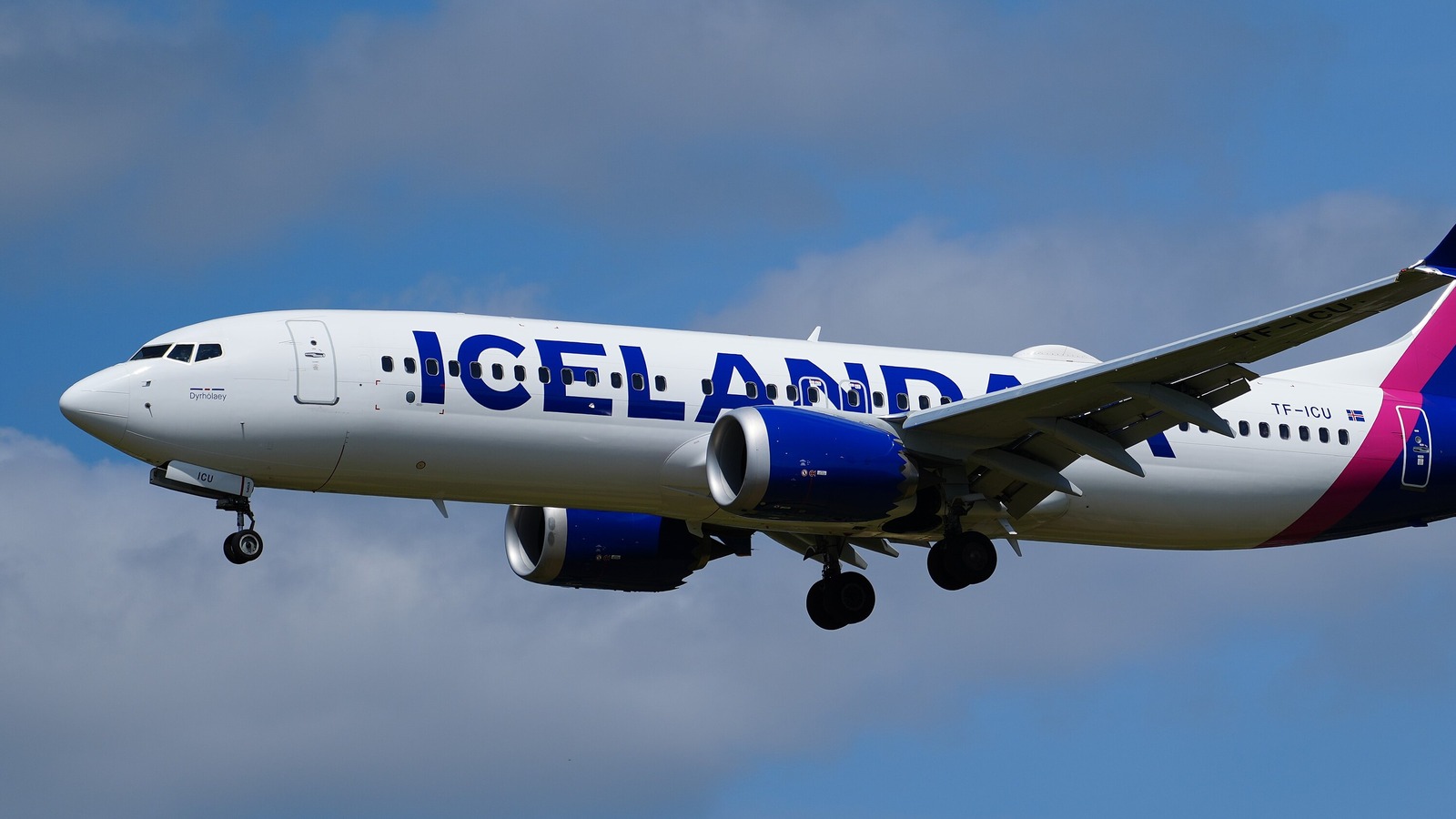 An Icelandair Boeing 737-8 MAX in white and blue livery with a magenta tail stripe, landing gear down, against a blue sky with clouds.