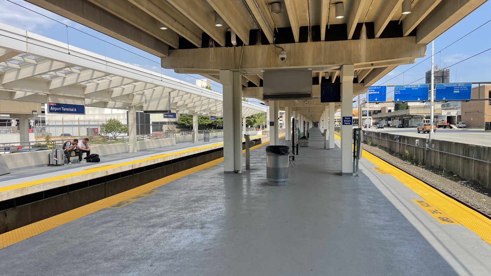 The regional rail platform at Philadelphia International Airport, with an Airport Terminal A nameboard, blue overhead signs directing passengers to Terminals B, C, D, E and F, and travellers waiting with suitcases