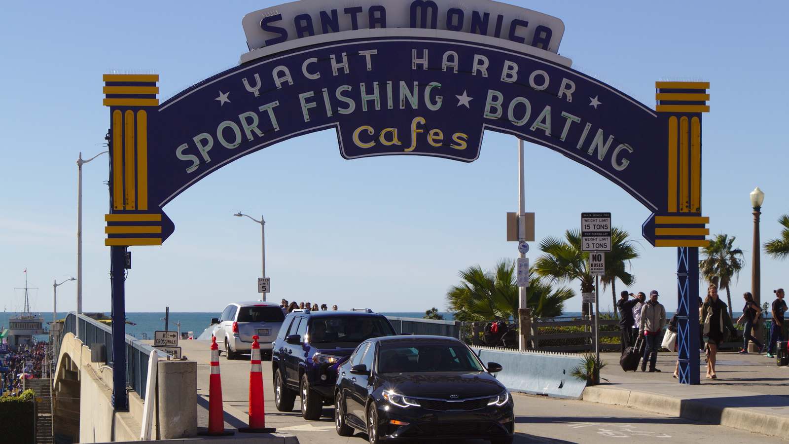 The Santa Monica Pier entrance arch over the bridge roadway, with cars driving under the sign and the ocean behind.