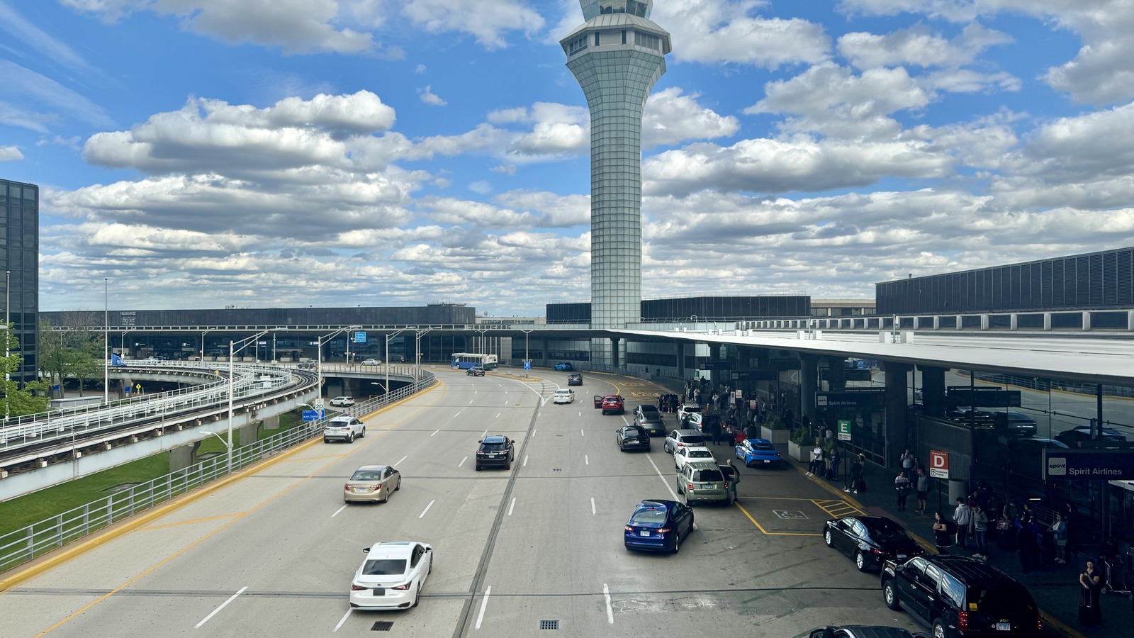 The control tower at Chicago O'Hare International Airport rising above the Terminal 2 and 3 kerbside roadway, with cars at the departures doors under a cloudy sky