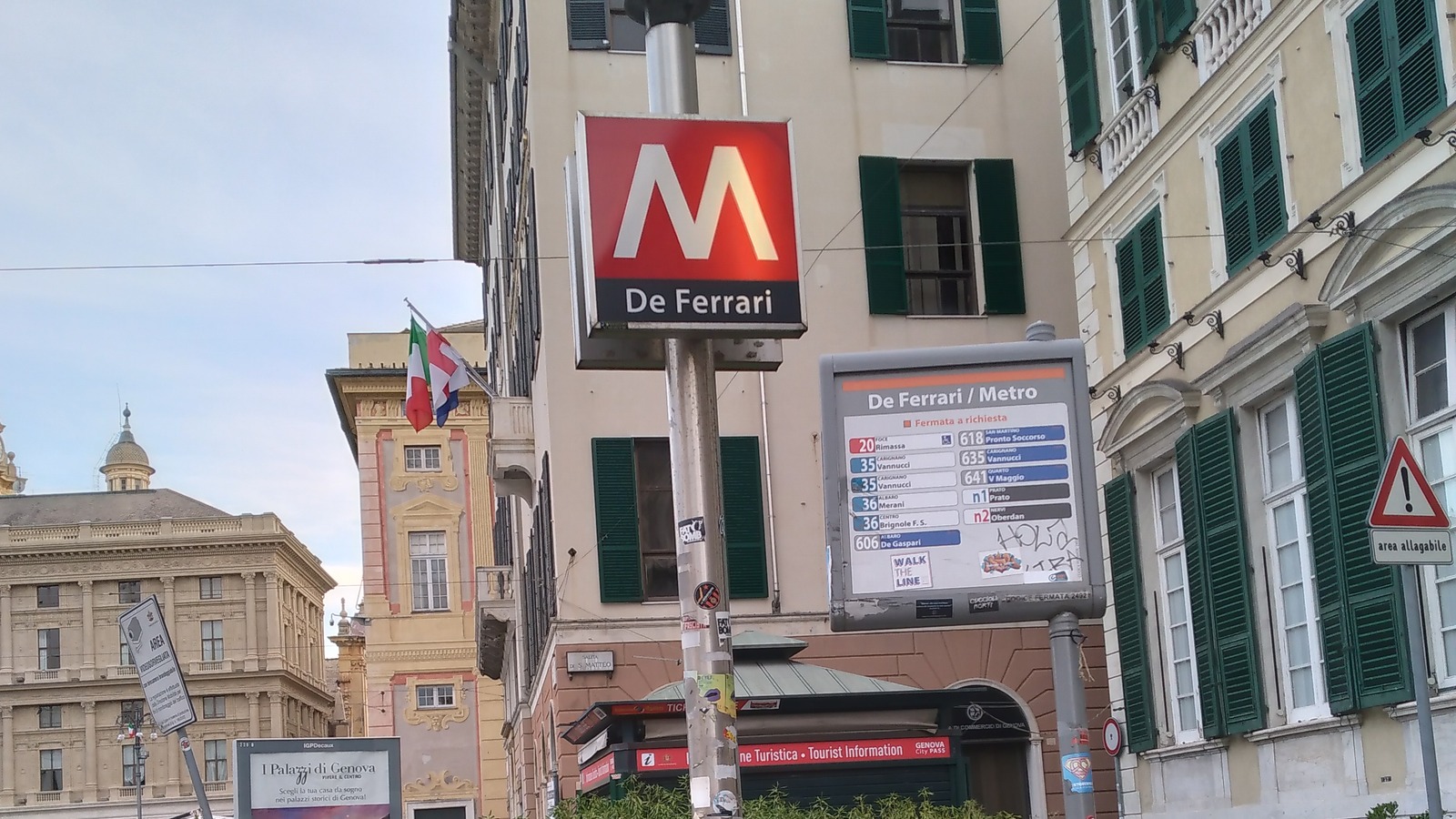 The De Ferrari metro entrance sign in central Genoa beside an AMT stop board listing bus routes