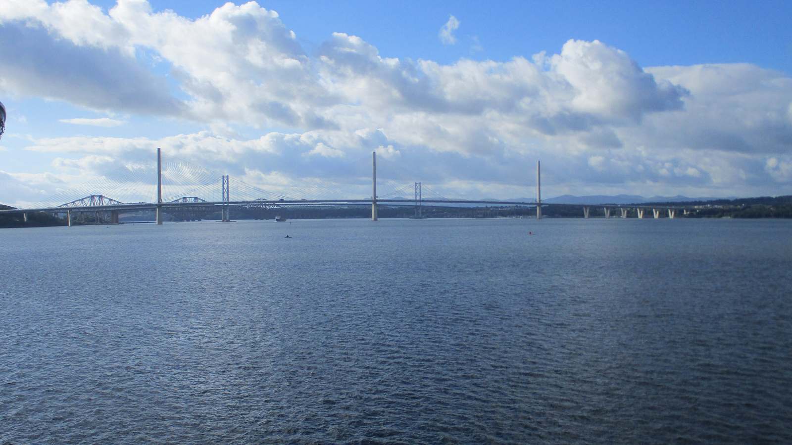 The three bridges over the Firth of Forth seen across the water from Rosyth, under a blue sky with white clouds