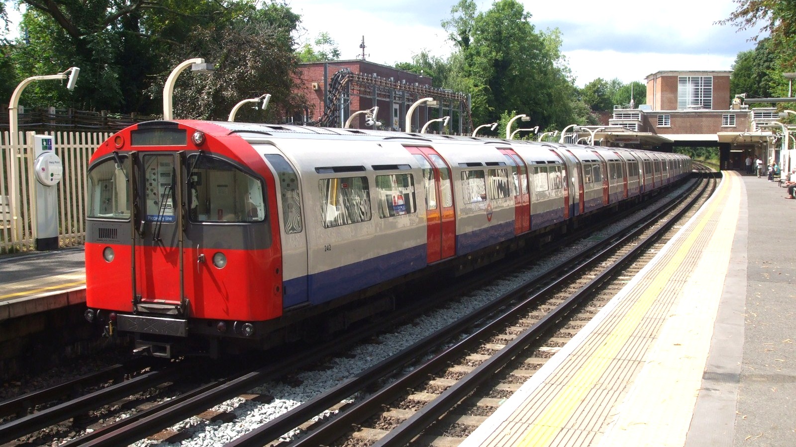 A red, white and blue Piccadilly line train of 1973 stock standing at the platform of Eastcote station in west London on a sunny day, with trees and the brick station building behind