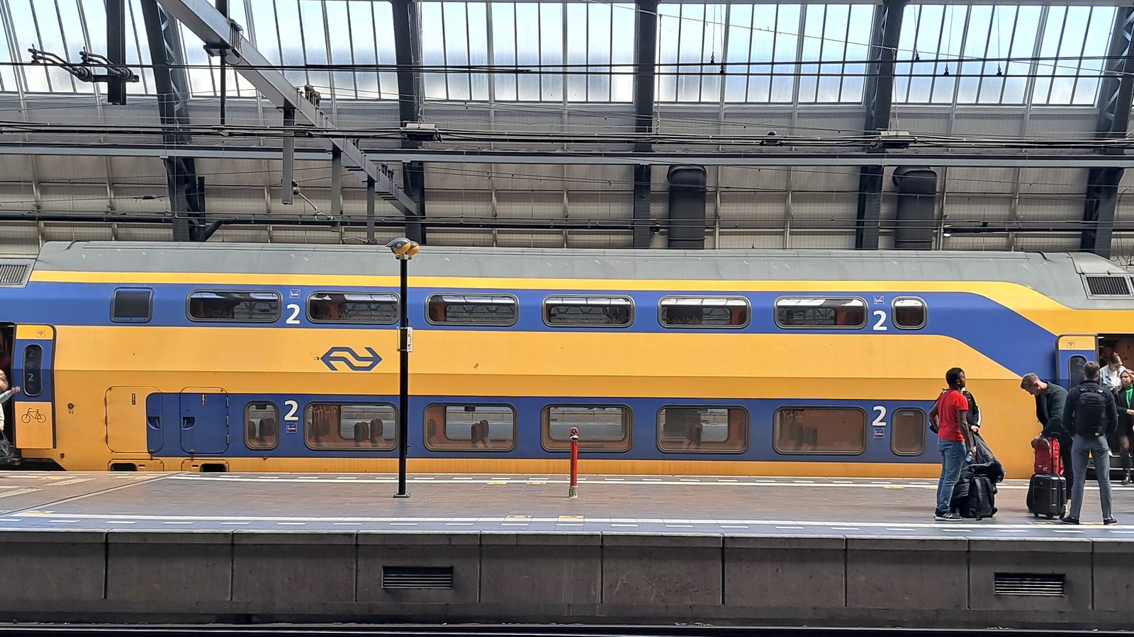 NS double-decker passenger train in yellow and blue livery at an Amsterdam Centraal platform with travelers boarding