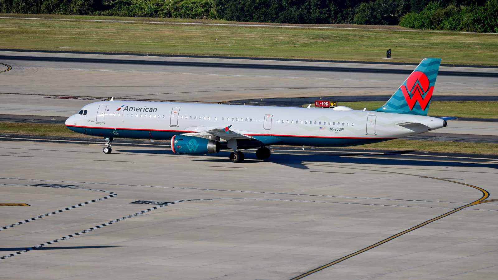 An American Airlines Airbus A321 in a retro tail livery taxiing on a sunlit taxiway, with grass and treeline behind it.
