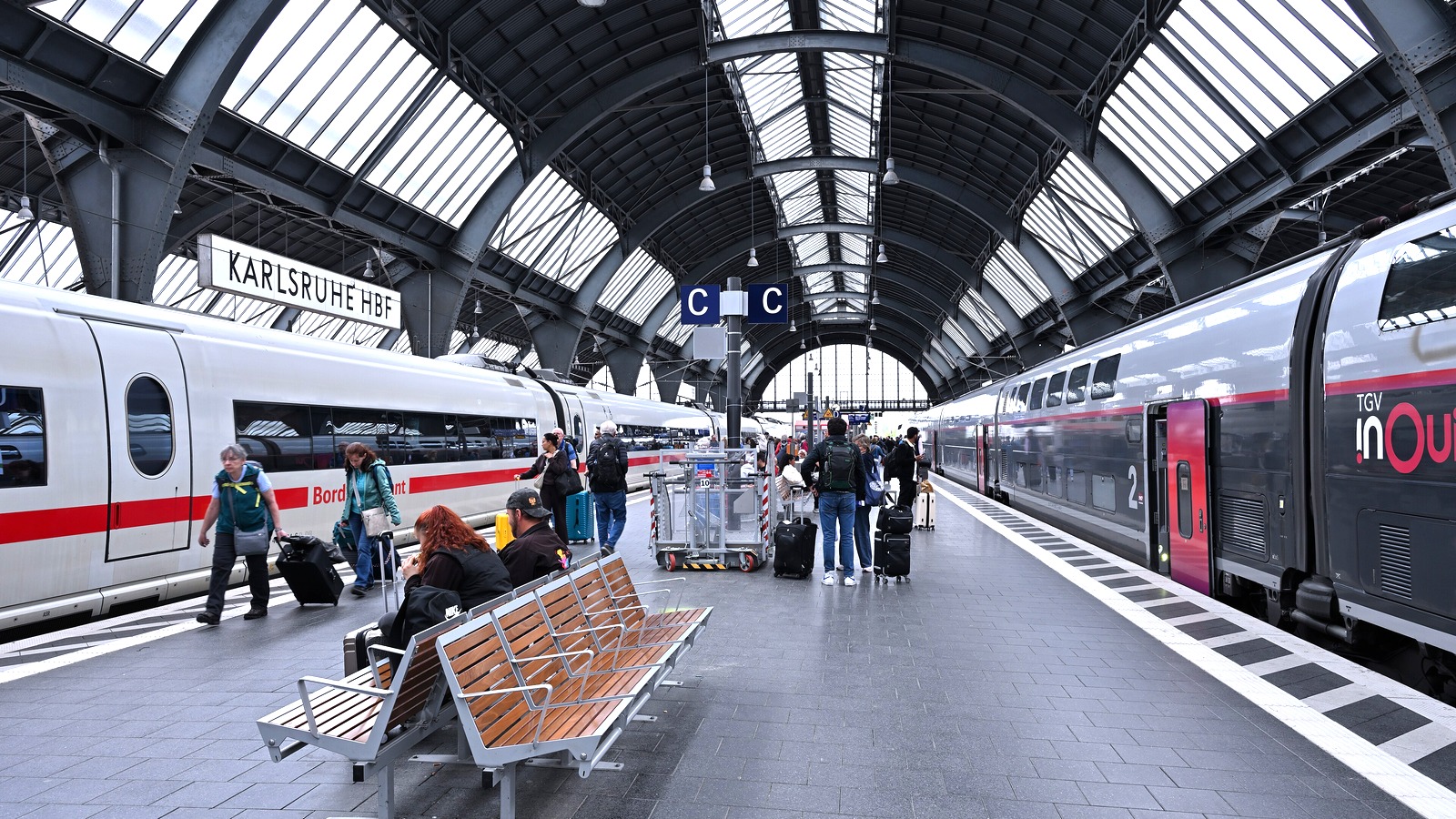 A German ICE train and a French TGV inOui stand at opposite platforms under the arched glass roof of Karlsruhe Hauptbahnhof, with passengers walking between them with wheeled luggage.