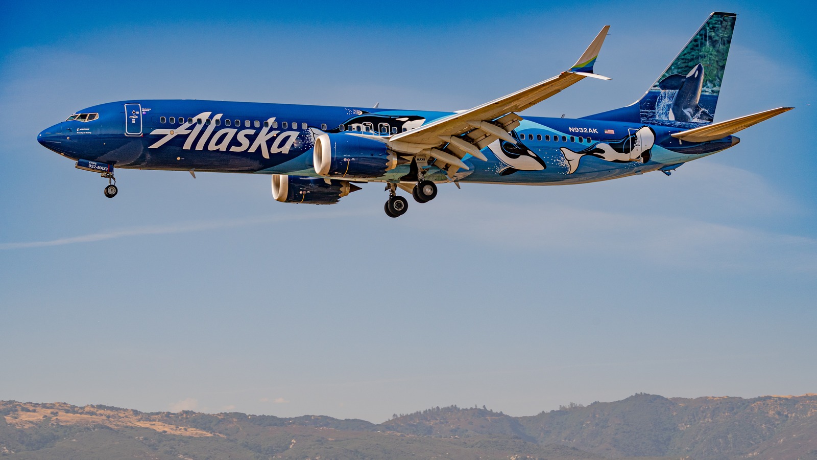 An Alaska Airlines Boeing 737 MAX 9 in the West Coast Wonders livery on approach, landing gear down, with hills below