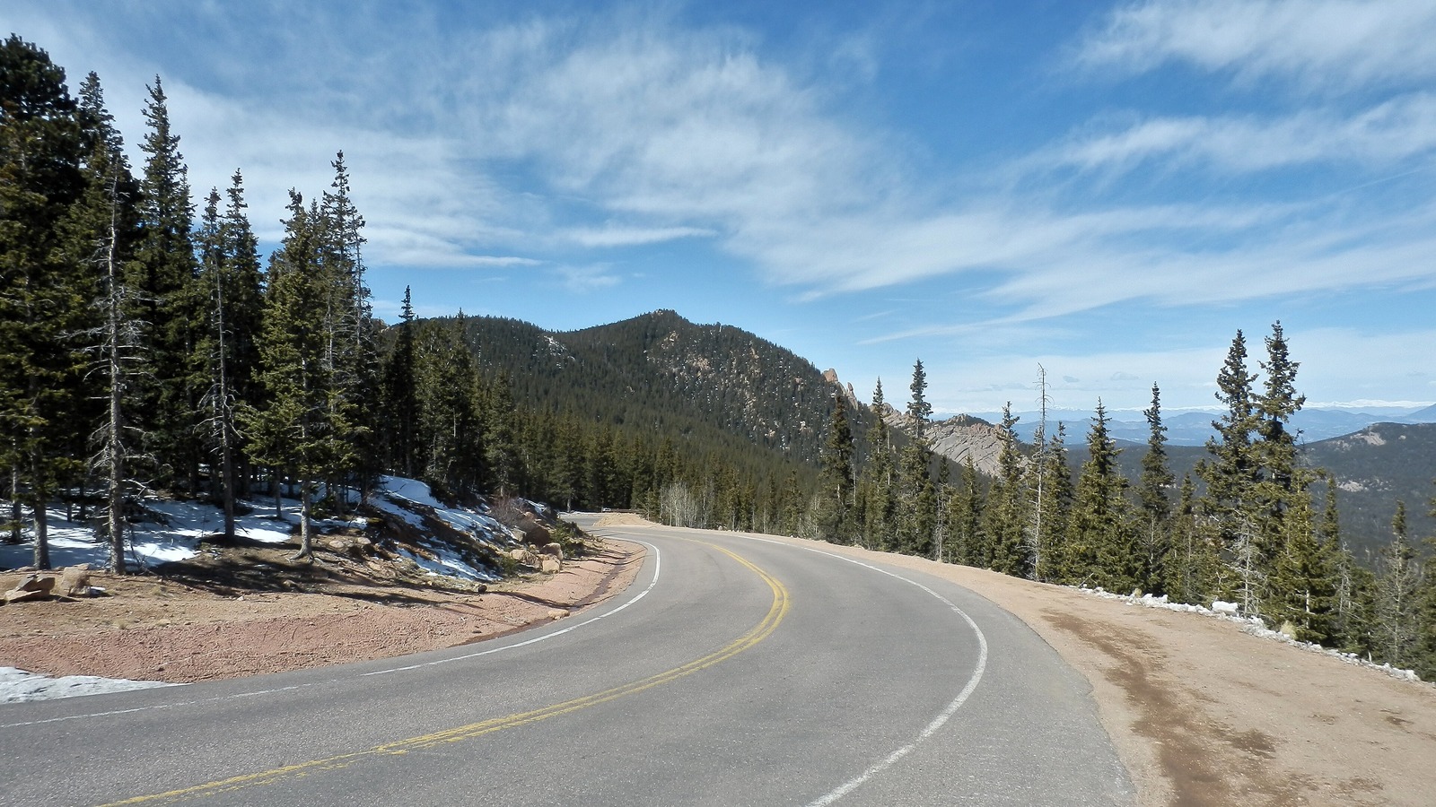 The Pikes Peak Highway curving right around a high country bend with a double yellow centre line, patches of old snow beside the shoulder and conifer forest falling away to distant ranges