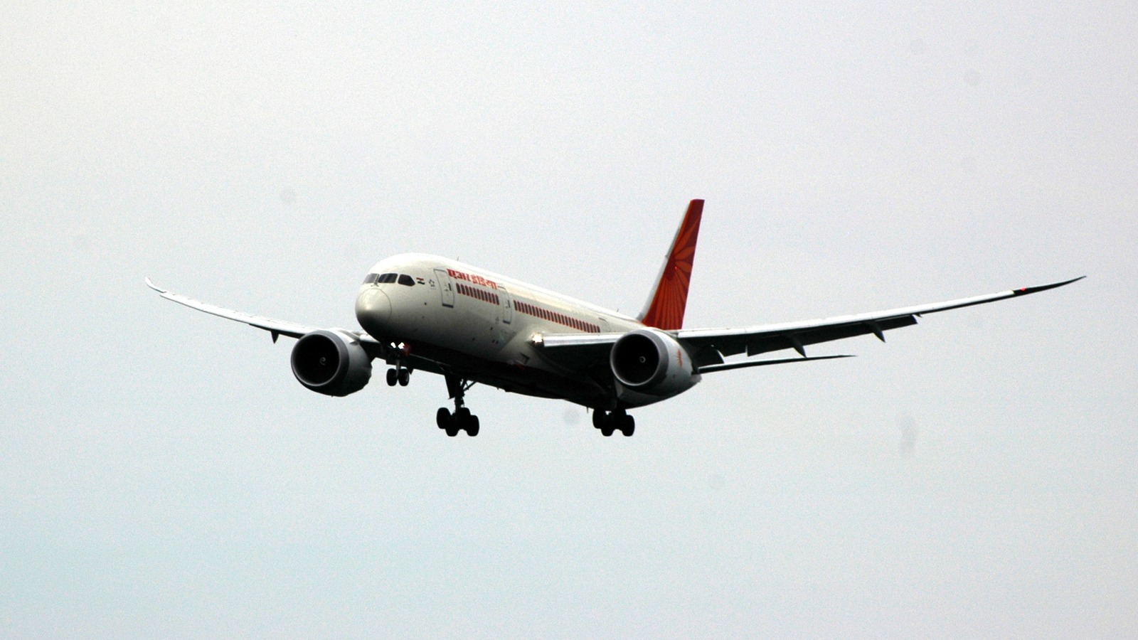 An Air India Boeing 787-8 Dreamliner with an orange tail on approach with landing gear down against a pale grey sky.