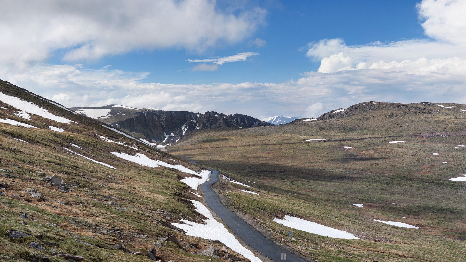 The narrow paved Mount Blue Sky road running above treeline across an alpine basin, with old snowbanks beside the asphalt, rocky tundra slopes either side and a jagged ridge and snow covered peaks behind.