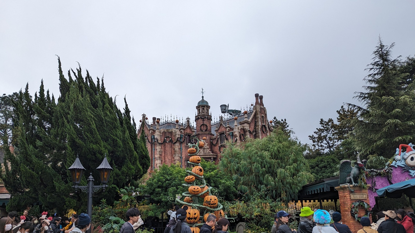 The Haunted Mansion at Tokyo Disneyland under its Holiday Nightmare overlay, with a spiral pumpkin tree, garlands and wreaths on the facade under an overcast sky