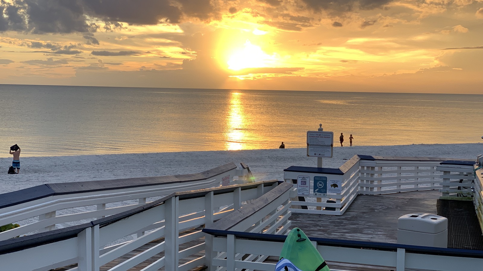 Sunset over the Gulf of Mexico from the Clam Pass Beach boardwalk in Naples, Florida, with white sand, a wooden walkway, and a kayak in the foreground.
