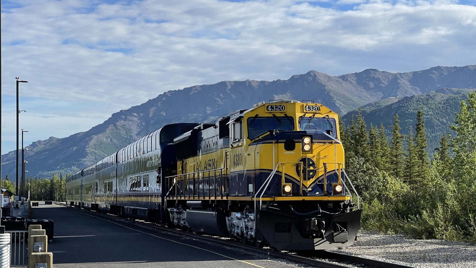 An Alaska Railroad passenger train in blue and gold livery standing at the Denali National Park depot platform, its dome cars stretching back along the track with forested mountains behind.