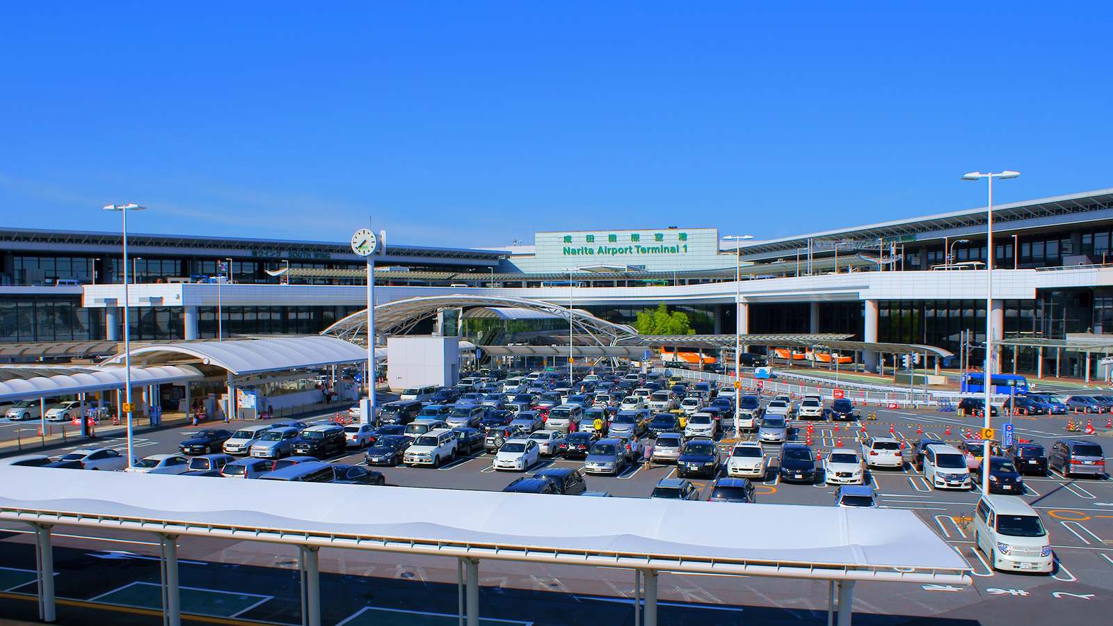 The front of Narita Airport Terminal 1 with its green sign, a large car park and bus shelters in the foreground under a clear blue sky.