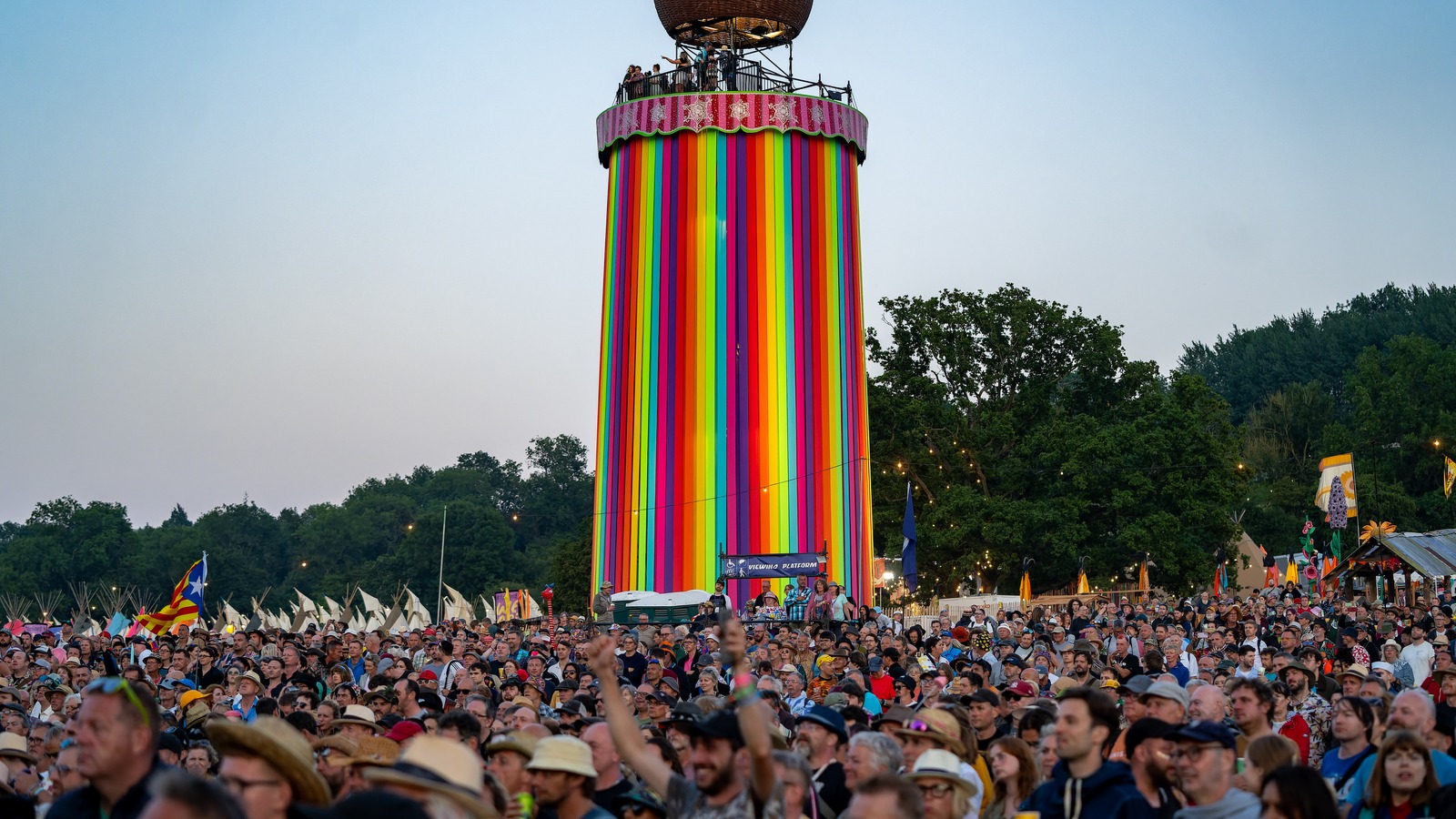 A dense crowd faces a tall, rainbow-striped tower at dusk during Glastonbury Festival, with trees and tent flags behind.