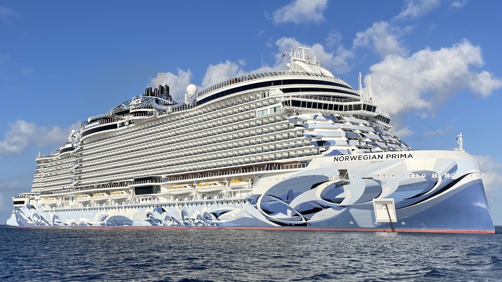 The Norwegian Cruise Line ship Norwegian Prima at anchor in calm blue water off George Town, Grand Cayman, with her name on the bow and the blue and white hull artwork visible