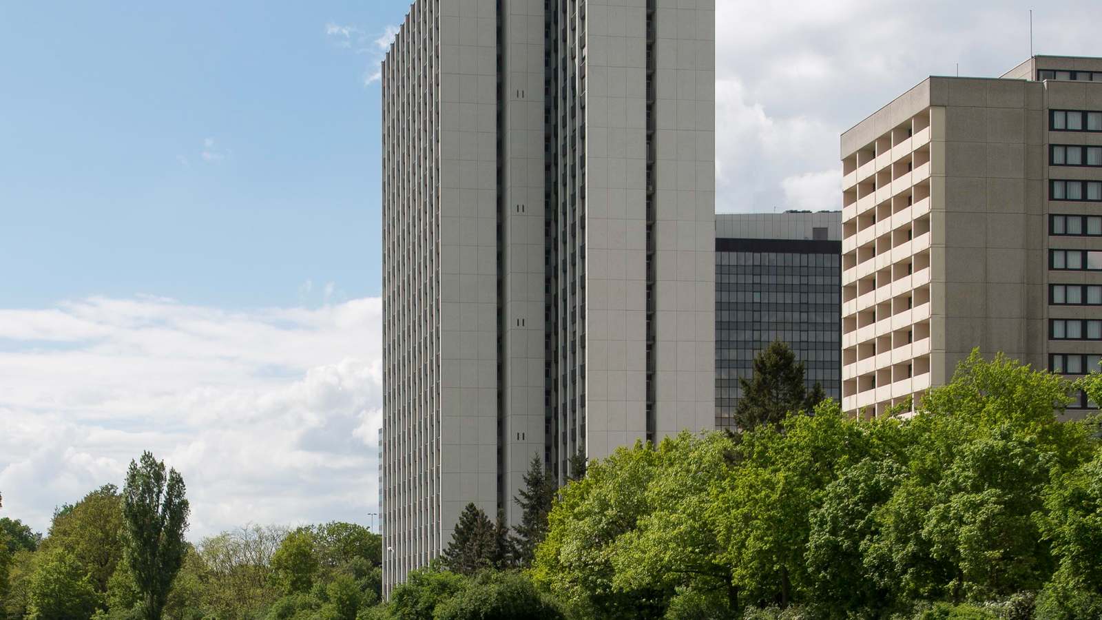 High rise buildings of the Arabella complex on Lyoner Strasse in Frankfurt Niederrad, behind a line of green trees under a partly cloudy sky.