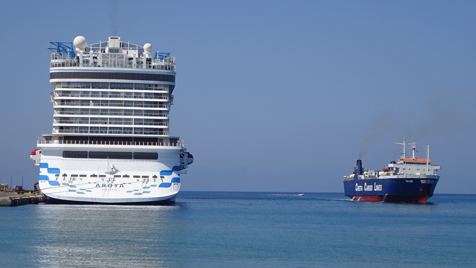 The stern of the cruise ship Aroya berthed at the Port of Rhodes in Greece, with AROYA and VALLETTA lettering visible on the hull and a cargo ship passing beyond
