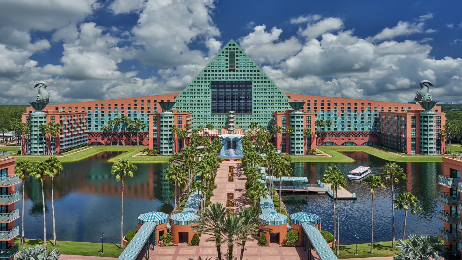 Walt Disney World Dolphin hotel exterior, a red-orange resort with a green pyramid roof, rooftop dolphin statues and a water fountain, seen across the resort lagoon and palm-lined walkway.