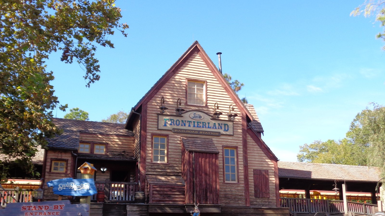 The Frontierland Railroad Station at Magic Kingdom photographed in December 2016, a timber building with a painted station sign under a clear sky, with an older Splash Mountain standby entrance sign at lower left