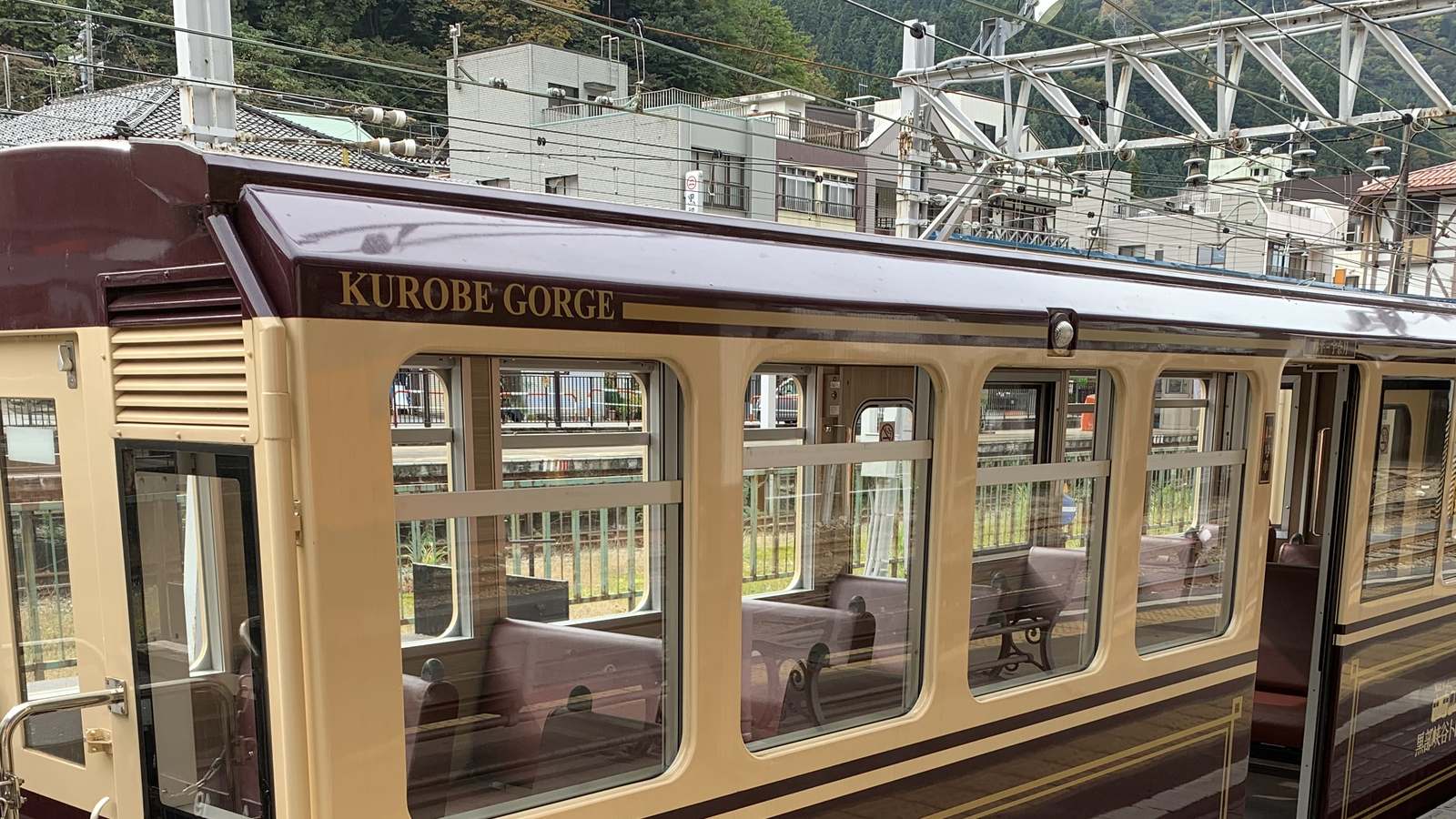 Maroon and cream Kurobe Gorge Railway passenger car with open windows and bench seats at Unazuki station, with buildings and wooded hills behind.