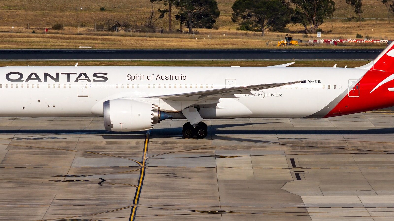 A Qantas Boeing 787-9 Dreamliner in white and red livery taxiing at Melbourne Airport with grassland behind.