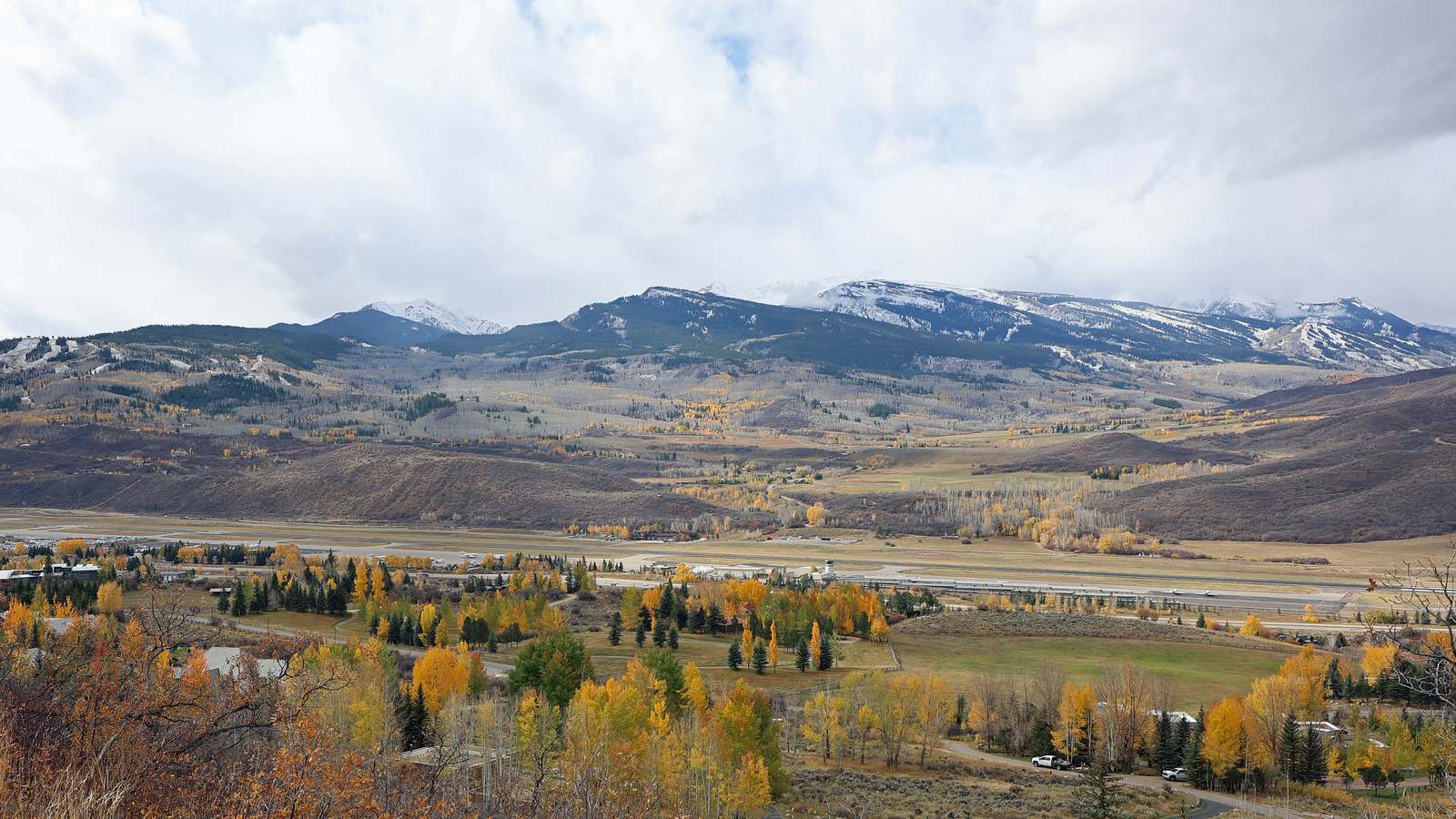 Aspen/Pitkin County Airport runway and terminal area in the valley below snow-dusted mountains and autumn aspens