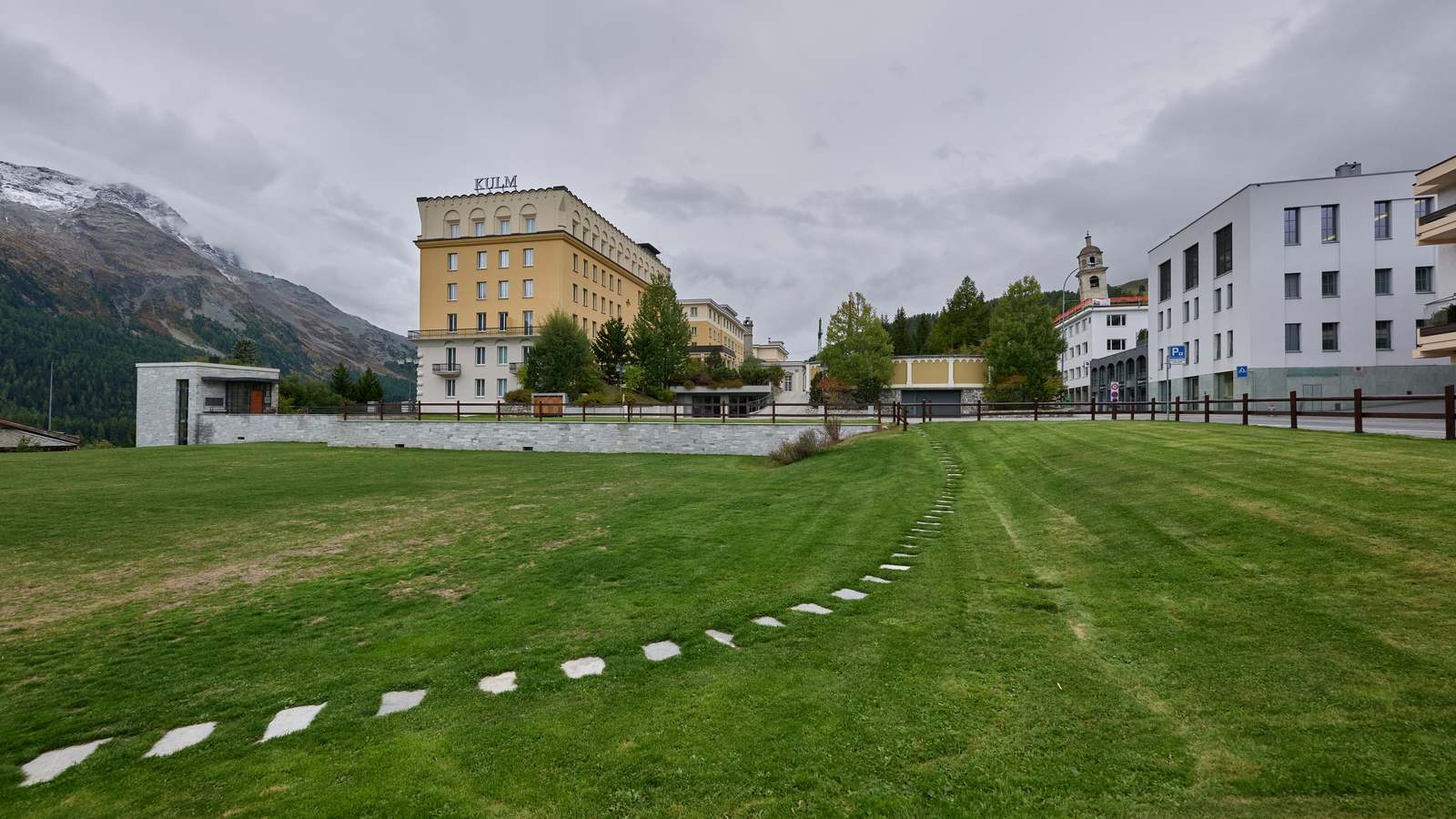 The yellow Kulm Hotel in St. Moritz, one of Europe's new Three Key hotels, beside a green lawn with mountains and cloud behind.