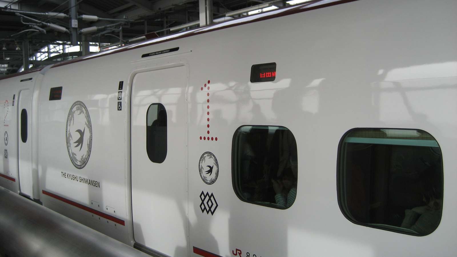 The side of a white Kyushu Shinkansen 800 series train at a station platform, showing the Kyushu Shinkansen emblem and car number.