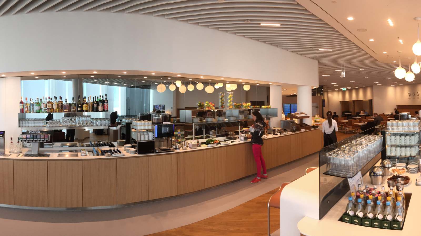 Buffet counter and bar inside the Lufthansa Senator Lounge at Munich Airport, with a few guests and staff and seating in the background.