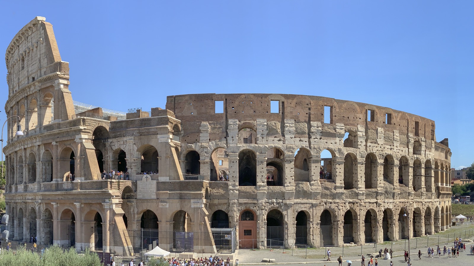 Exterior of the Colosseum in Rome under a clear sky, with visitors on the surrounding pavement