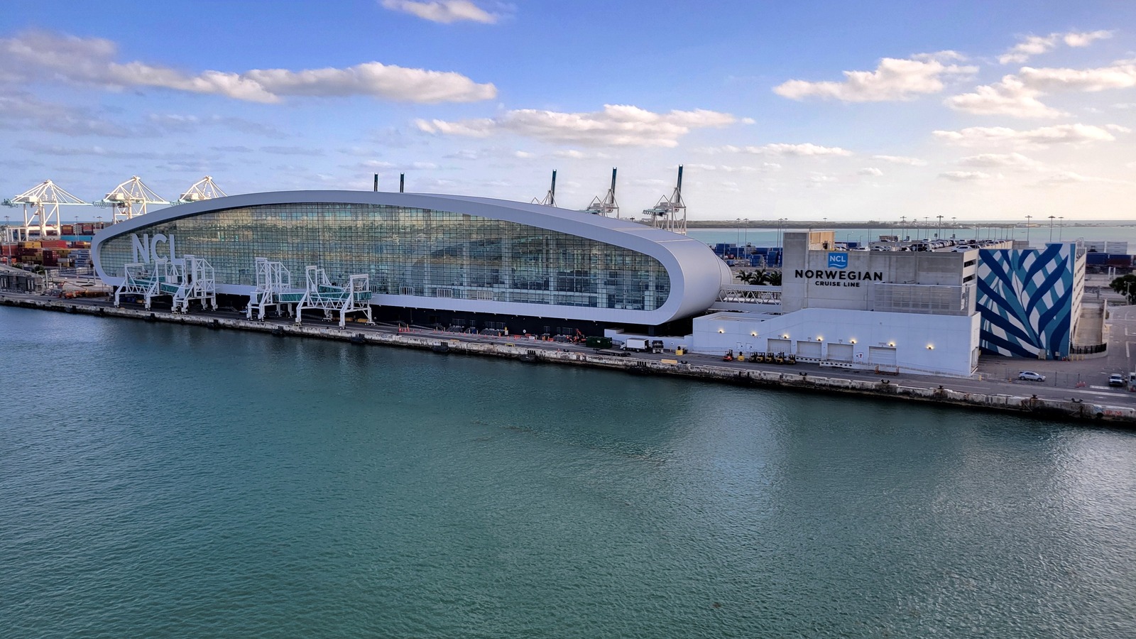 Cruise terminal building on Dodge Island at PortMiami seen from the water, with cargo cranes and the port bridge behind it