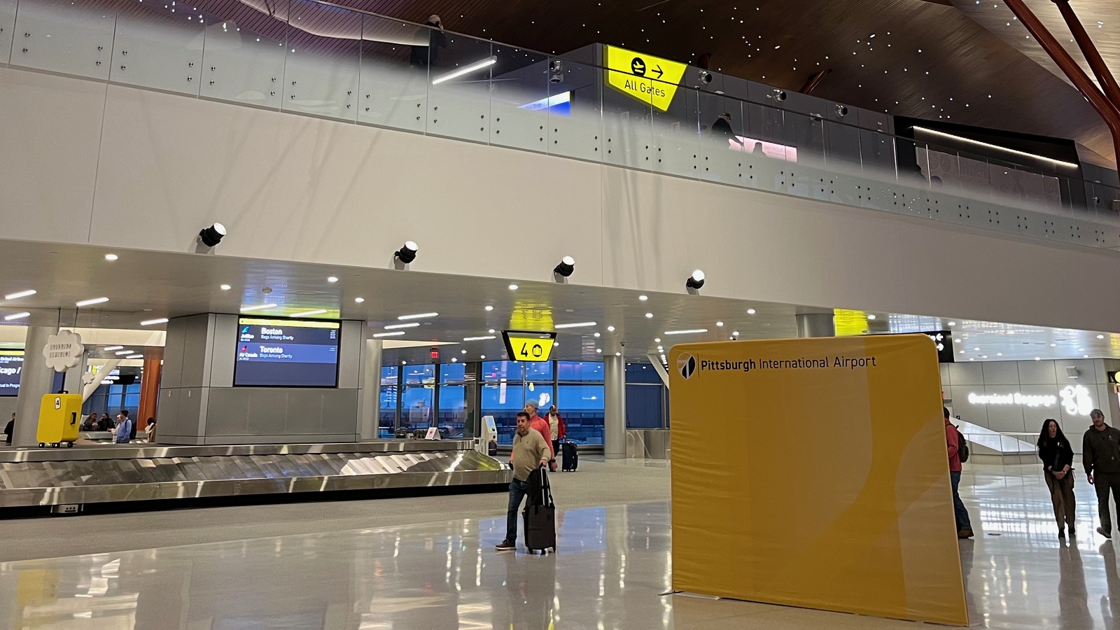 Baggage claim hall inside the new Pittsburgh International Airport terminal, with a Pittsburgh International Airport sign and arriving passengers collecting bags.