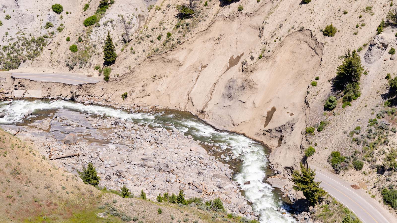 An aerial view of the Gardner River rushing past a washed out section of Yellowstone's North Entrance Road in the Gardner Canyon after the 2022 flood, with intact road on either side.
